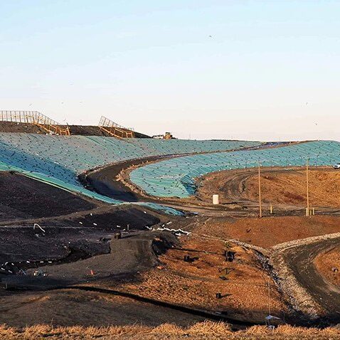 A large landfill site with sloped areas covered in green tarps or liners, surrounded by barren soil and gravel paths under a clear sky. Some fencing and utility poles are visible in the distance.