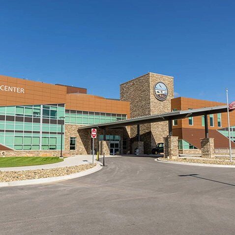 A modern building labeled Oyate Health Center with large windows, a stone facade, two flagpoles with American and South Dakota flags, and a covered entrance under a clear blue sky.